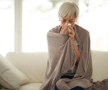 Elderly woman wrapped in a blanket, sitting on a couch, blowing her nose with a tissue.