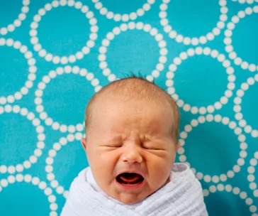 A crying baby swaddled in a white blanket against a teal background with white circular patterns.