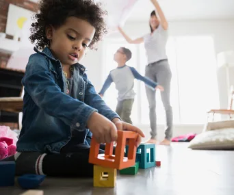A toddler plays with colorful blocks on the floor while two adults play in the background.