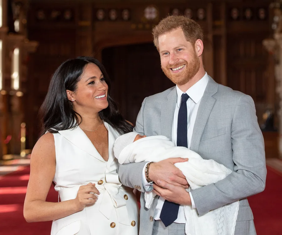 A smiling couple holding their newborn wrapped in a white blanket in an ornate room.