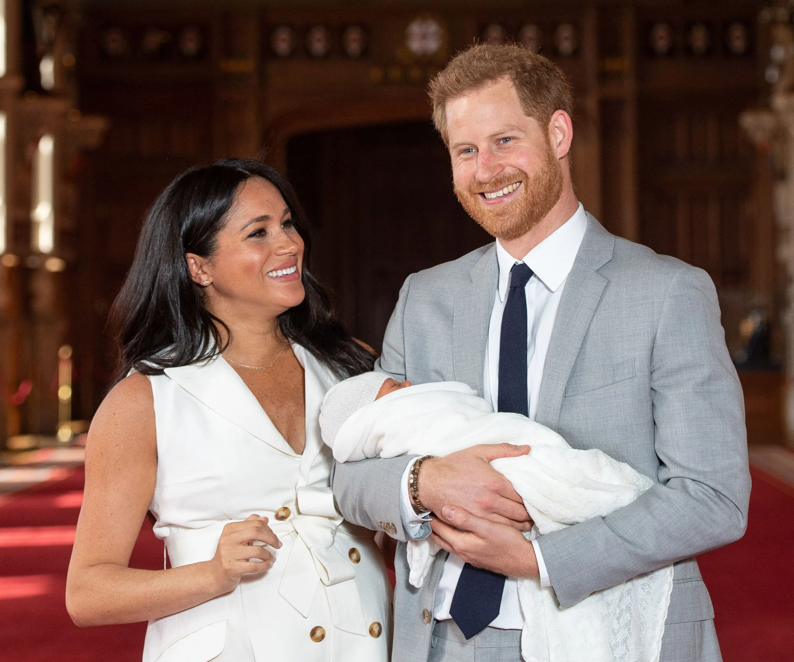 A smiling couple holding their newborn wrapped in a white blanket in an ornate room.