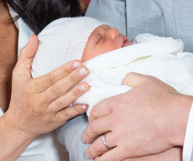 Newborn baby wrapped in a white blanket, wearing a knit cap, cradled by two adults.