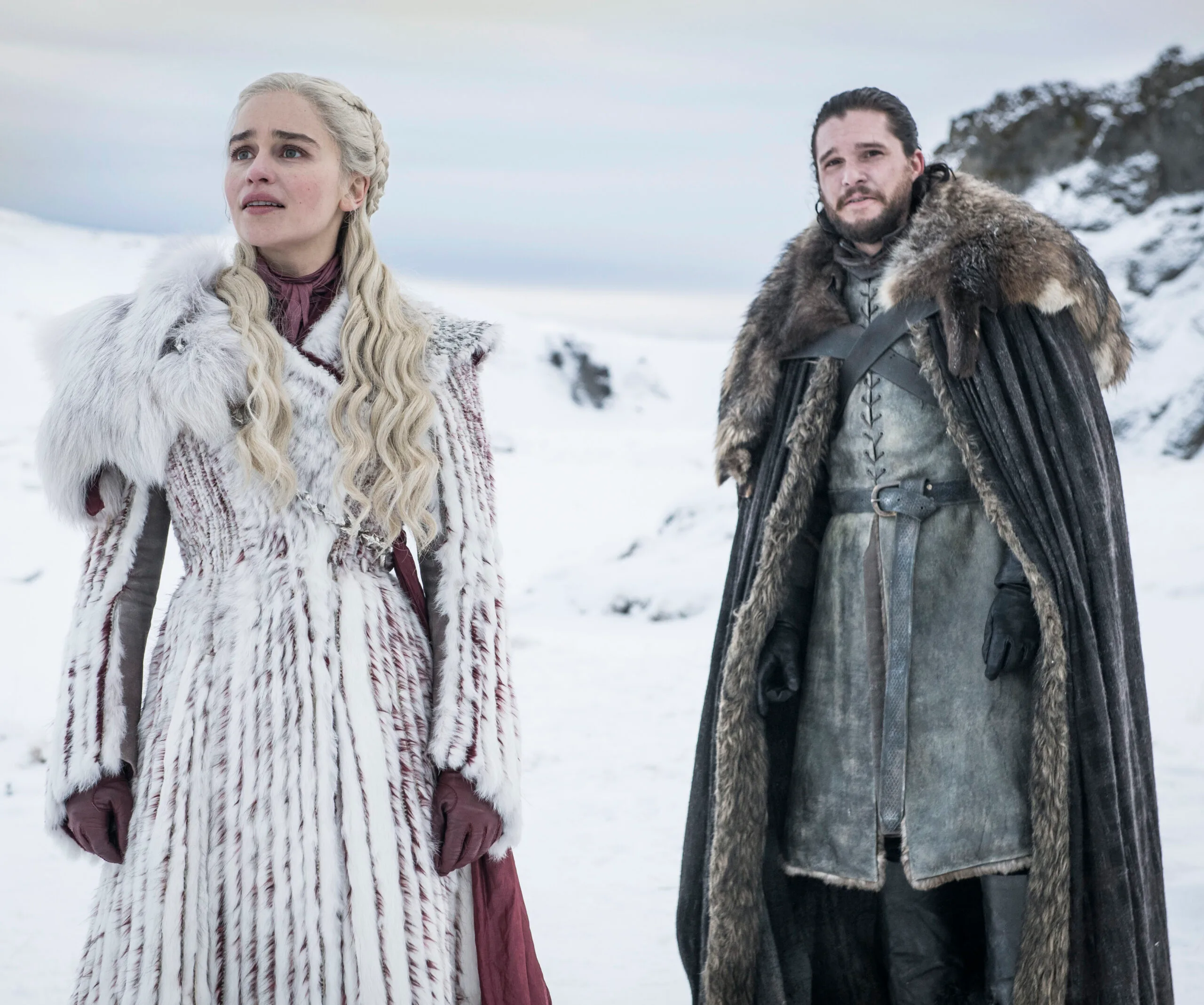 Daenerys Targaryen and Jon Snow stand in snowy landscape wearing fur-trimmed coats.