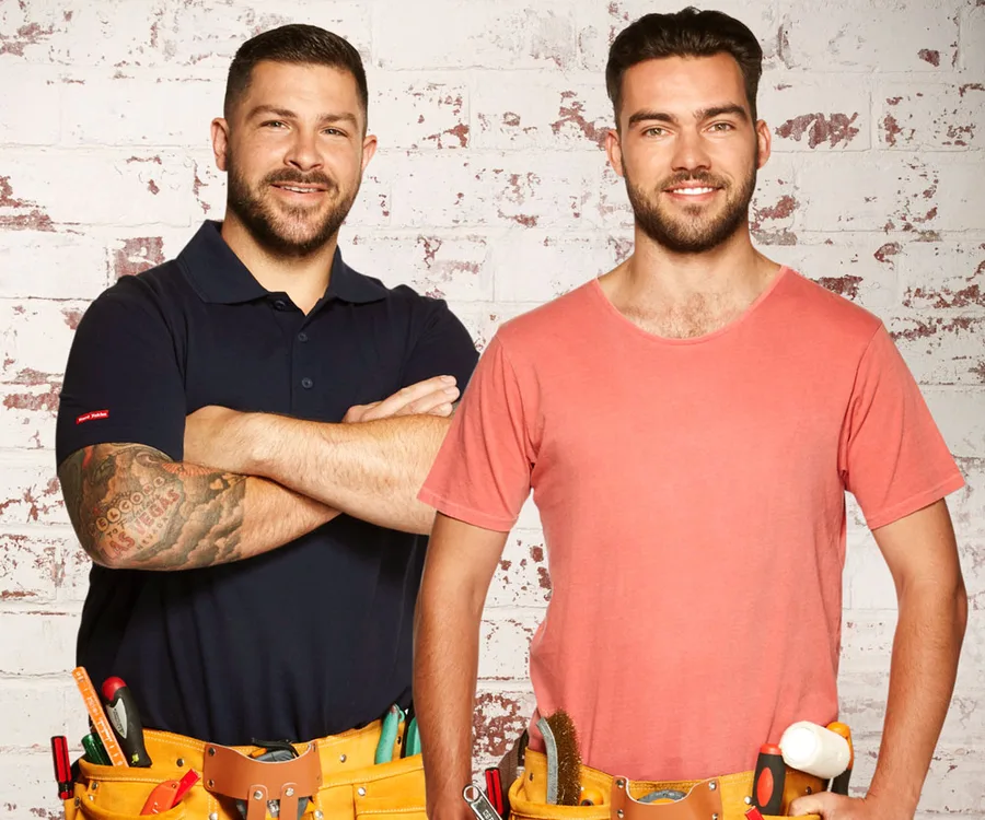 Two men smiling, wearing tool belts, standing against a brick wall. One has a tattoo, both look ready for house work.