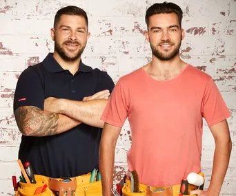 Two men smiling, wearing tool belts, standing against a brick wall. One has a tattoo, both look ready for house work.