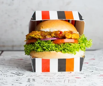 Vegan burger with lettuce, tomato, and patty in a colorful striped box on a patterned surface.