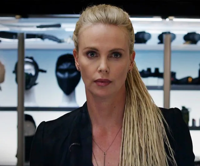 Cipher from "Fast & Furious" series with long blond hair, wearing a black outfit, stands in front of display shelves.
