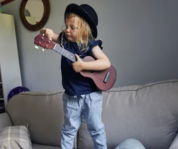 Toddler standing on a couch wearing a black hat, playing a small guitar.