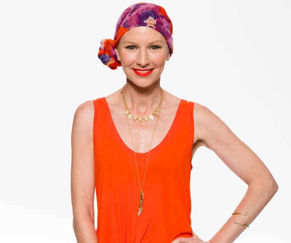 A woman in an orange top, wearing a colorful headscarf, smiles against a plain background.