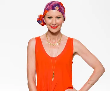 A woman in an orange top, wearing a colorful headscarf, smiles against a plain background.