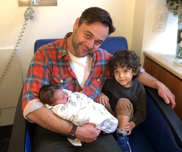 Father holding a newborn in a hospital, with a young child sitting beside him.