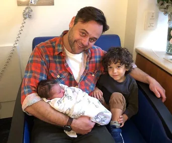 Father holding a newborn in a hospital, with a young child sitting beside him.