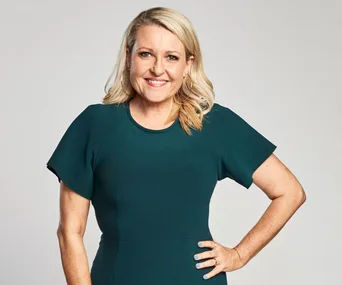Blonde woman in a green dress smiling, hand on hip, against a neutral background.