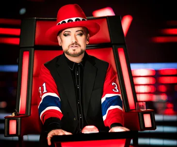 Red and black dressed coach in a hat sits on The Voice Australia's set with colorful lighting.