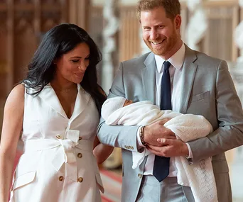 A couple smiling while holding a newborn wrapped in a white blanket inside a grand hall.