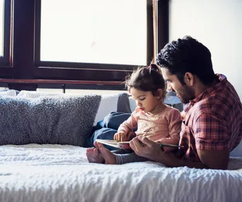 Father and toddler reading a book together on a cozy bed with soft pillows.