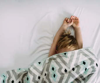 Child sleeping under patterned blanket, arms stretched above head.