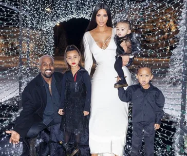 Family portrait with four people standing and one person holding a child, surrounded by twinkling lights.