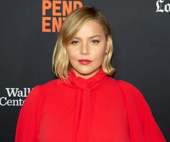 Abbie Cornish in a red dress at a premiere event, standing against a black backdrop with logo text.