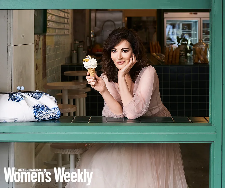 Woman in a pink dress holding an ice cream cone, smiling through a cafe window, with a blue and white bag beside her.