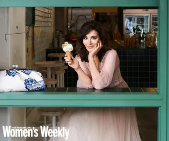 Woman in a pink dress holding an ice cream cone, smiling through a cafe window, with a blue and white bag beside her.