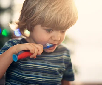 A toddler brushing teeth with a red and blue toothbrush, wearing a striped shirt, in soft natural light.