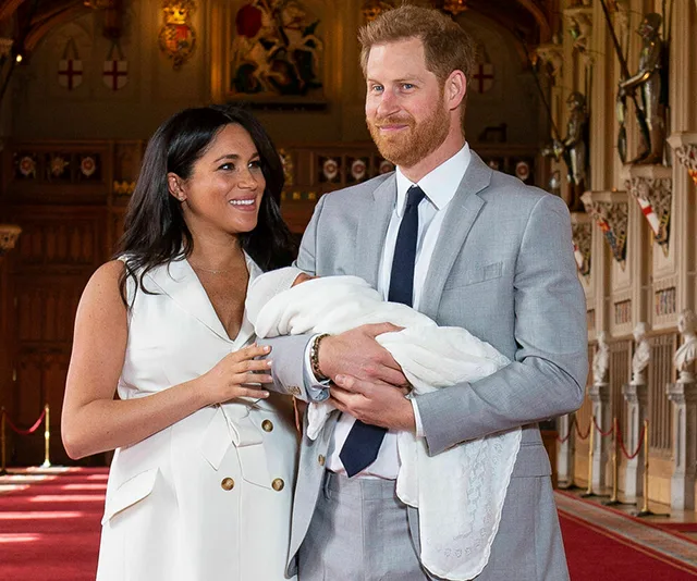 A couple smiling joyfully with a newborn baby wrapped in a white blanket, standing in an ornate hall.