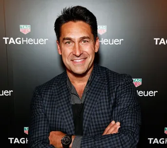 I don’t know who this is, but a person smiles with arms crossed at a TAG Heuer event backdrop, wearing a checked blazer.
