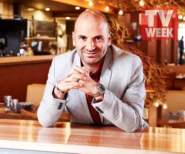 Man in a gray suit smiling with folded hands in a restaurant setting, TV WEEK logo in the corner.