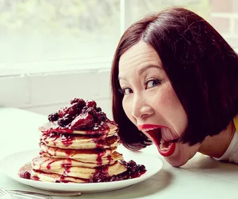 Woman playfully posing with a stack of berry-topped pancakes on a table near a window.