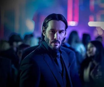 A man in a suit stands in a dimly lit, crowded room with colorful lights in the background, looking intently at the camera.