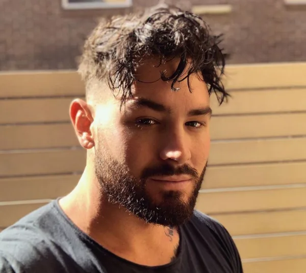 A man with a beard and short, curly hair takes a selfie outdoors, wearing a black shirt, with a fence in the background.