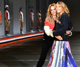 Two women embrace at a fashion event runway, with models lined up in background.