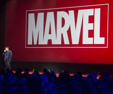 Man on stage with a microphone, large Marvel logo displayed on screen, audience in foreground.