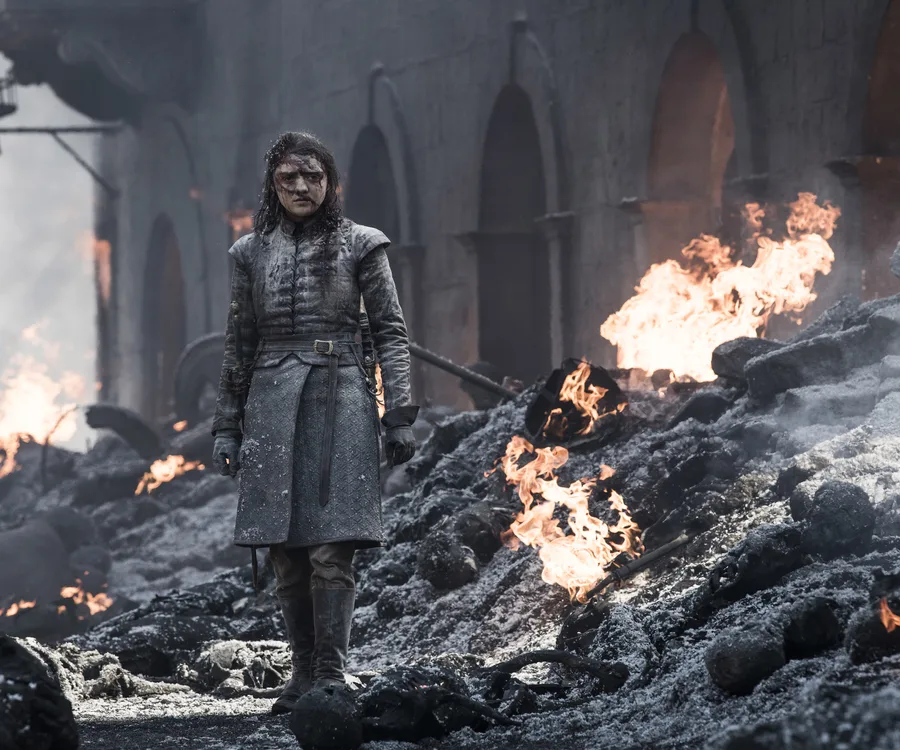 Arya Stark stands amidst ruins and flames in a battle-torn King's Landing.