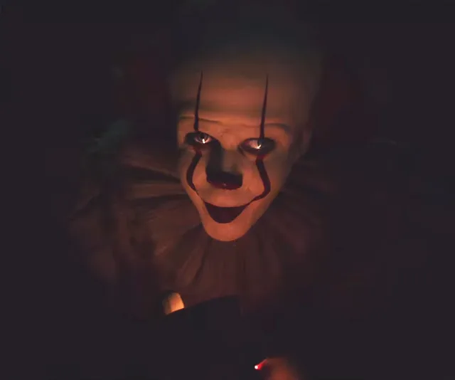 Pennywise the clown, with glowing eyes, looks menacingly in the dark from "It Chapter Two" movie scene.
