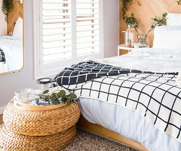 Cozy bedroom with a grid-patterned blanket, woven basket table, eucalyptus leaves, and natural light through shutters.