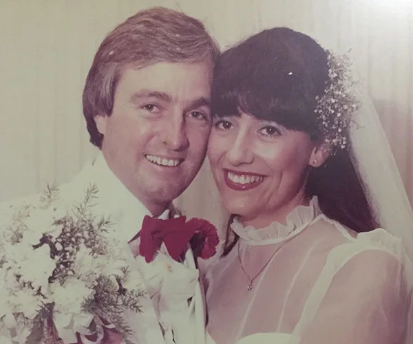 A couple on their wedding day, the bride in a white dress and veil, holding flowers, both smiling.
