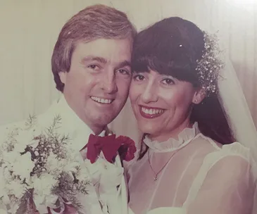 A couple on their wedding day, the bride in a white dress and veil, holding flowers, both smiling.