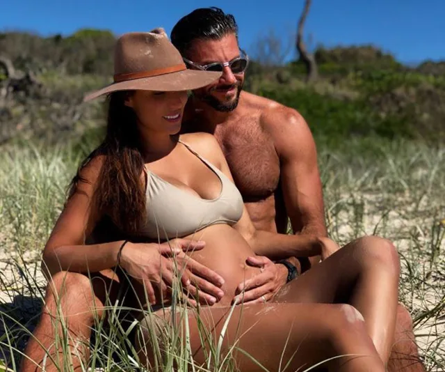 Couple sitting on grass at the beach, woman in bikini and hat cradling baby bump, man behind her in sunglasses.