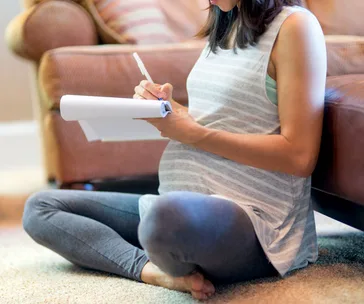Pregnant woman sitting on floor, writing on notepad, in casual attire.