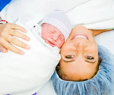 Mother holding swaddled newborn baby, both smiling after a cesarean delivery.