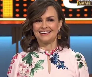 A woman with wavy hair, wearing a floral dress with a keyhole neckline, smiling on a game show set.