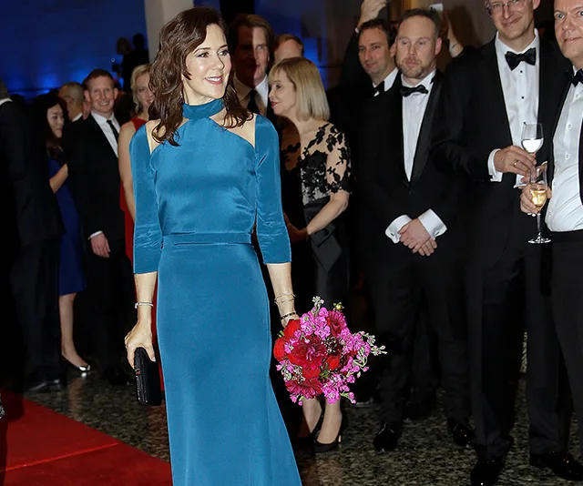 Elegant woman in blue gown holding flowers at formal event with attendees in background.