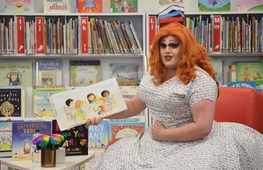 Drag queen with orange hair reads a children's book at a library, surrounded by colorful books on shelves.