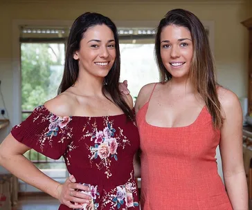 Two women smiling indoors; one in a floral dress, and the other in an orange dress.