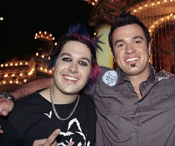 Two men posing together, one with dyed hair and piercings, in a lively setting with bright lights in the background.