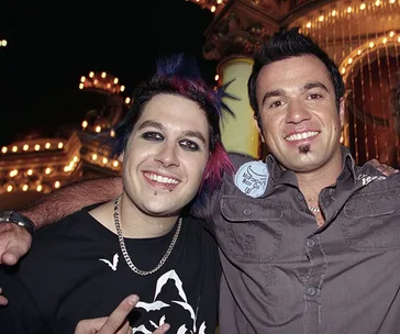 Two men posing together, one with dyed hair and piercings, in a lively setting with bright lights in the background.