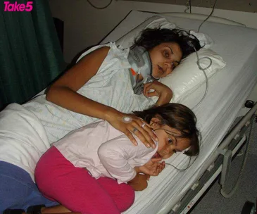 A woman with a neck brace lies in a hospital bed, embracing a child resting on her side.