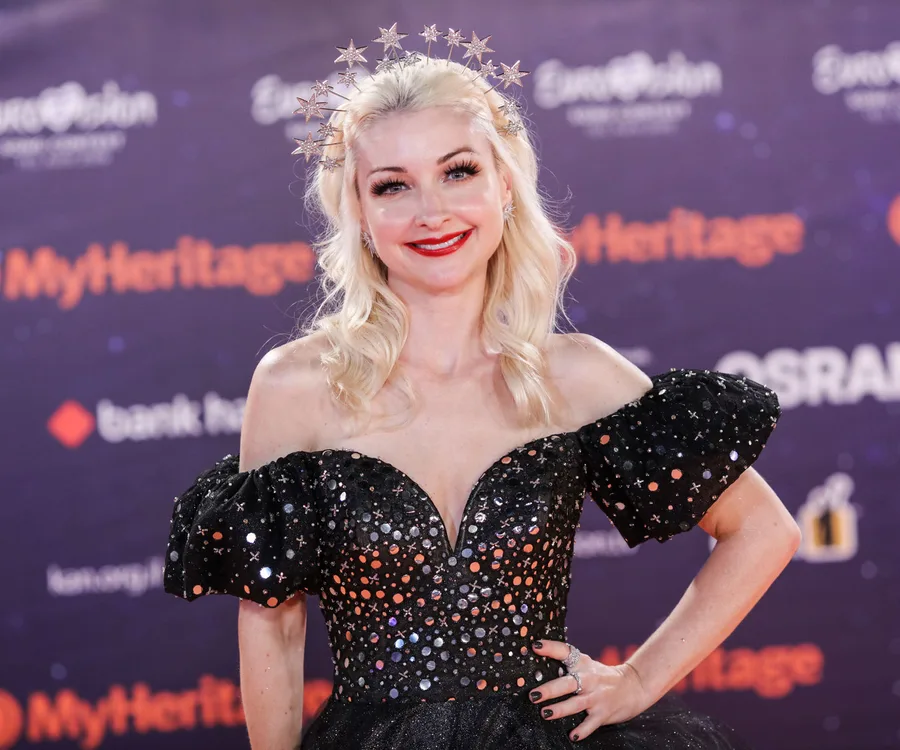 A woman in a sparkling off-shoulder dress and star-themed headpiece smiles on the Eurovision red carpet.